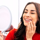 A woman wearing a red blouse admiring her new smile in the mirror after receiving her dental implant